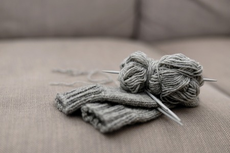 Grått garnnystan och stickade plagg med stickor ligger på en beige soffa.