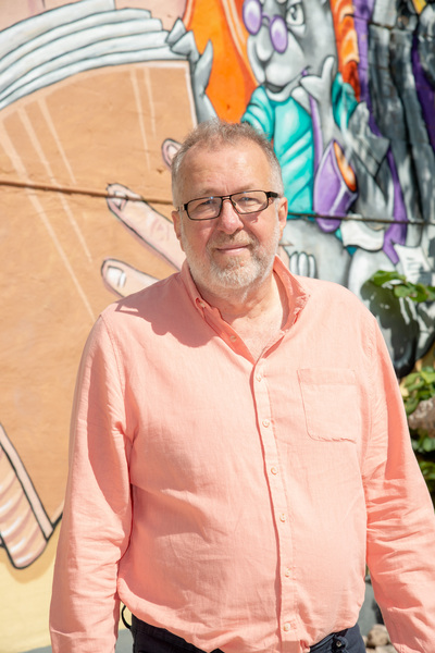 Man i ljusrosa skjorta står framför en färgglad väggmålning utomhus.