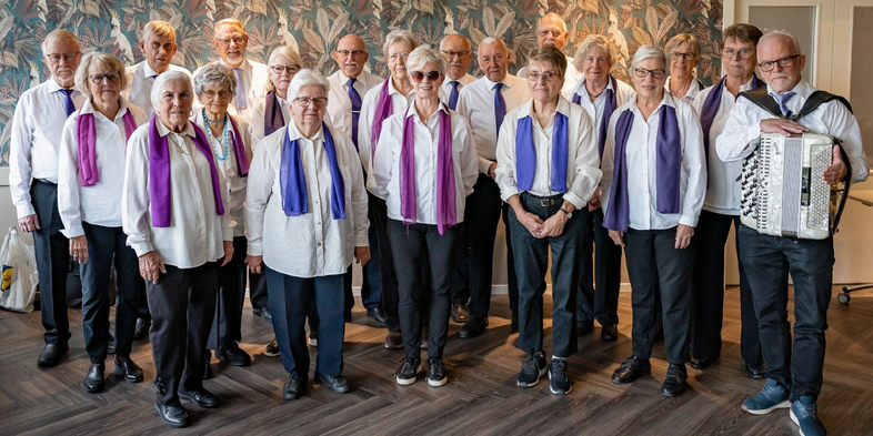 En grupp äldre personer står tillsammans inomhus, klädda i vita skjortor och färgade halsdukar. En person håller ett dragspel.