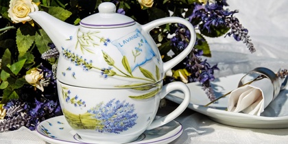 En tekanna och kopp med lavendelmotiv står på ett fat, omgiven av blommor och en servett på tallrik.