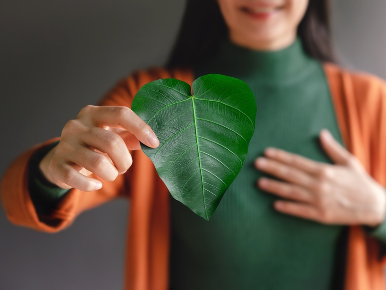 Kvinna håller ett grönt hjärtformat blad framför bröstet, klädd i grön tröja och orange kofta.