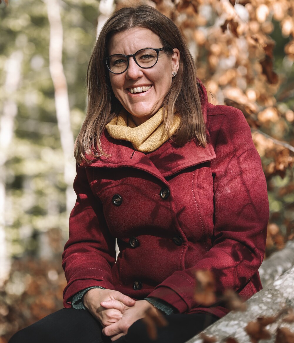 Kvinna med glasögon och röd kappa sitter utomhus bland höstlöv och ler mot kameran.