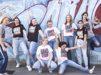 En grupp unga personer poserar glatt framför en graffitivägg, alla bär t-shirts med texten 'DANS STUDION'.