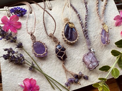 Fem handgjorda halsband med stenar och makramé ligger på ett papper, omgivna av lila och rosa blommor.