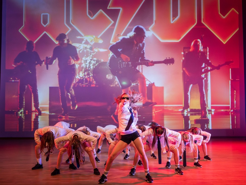 En dansgrupp uppträder på scen framför en stor bakgrundsbild av rockbandet AC/DC.