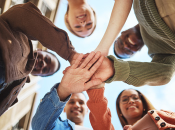 Fem personer står i en cirkel och lägger sina händer ovanpå varandra, sedda underifrån mot himlen.