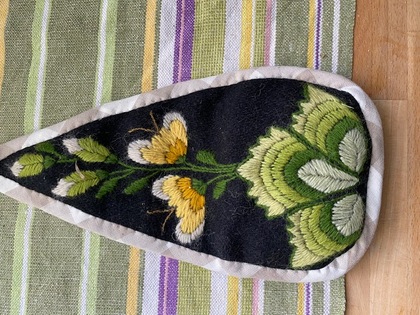 Broderad tygbit med blommotiv i grönt och gult på svart bakgrund, liggande på randig duk.