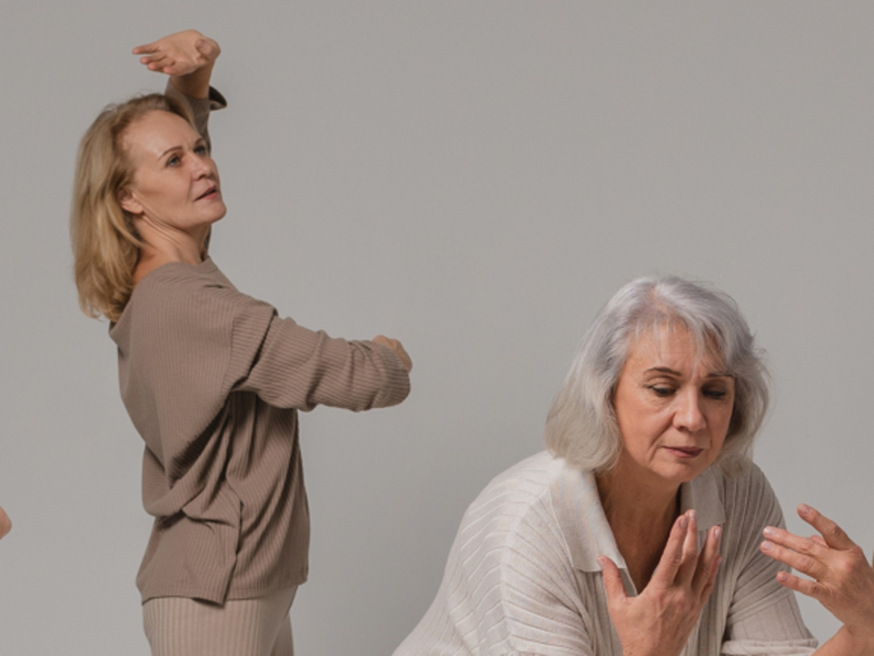 Fyra kvinnor i olika åldrar dansar barfota i mjuka, neutrala kläder mot en ljus bakgrund.