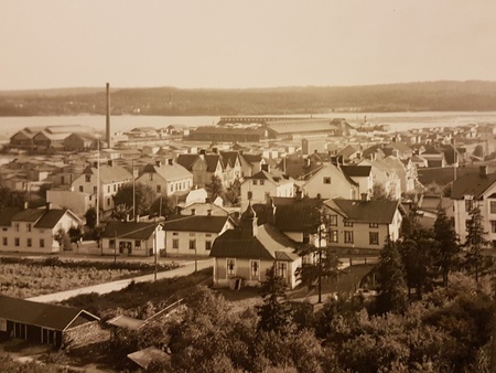 Gammalt svartvitt sepiafärgat foto över en småstad med trähus, fabriker och vatten i bakgrunden.