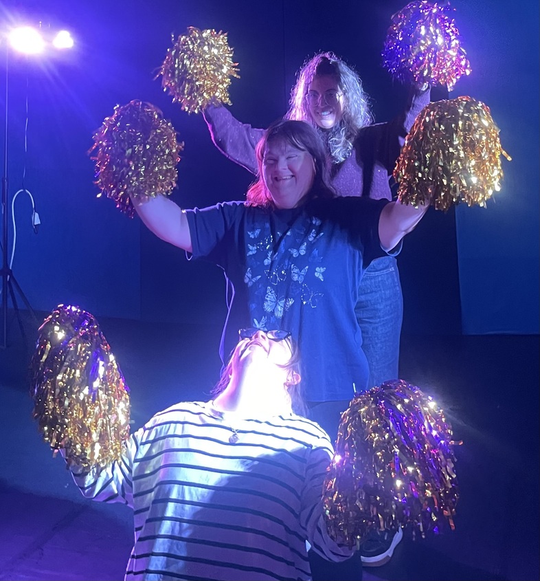 Tre personer står i rad och håller upp glittriga pompoms under stark scenbelysning.