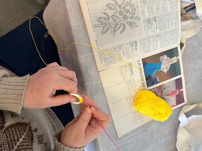 Person virkar med gult garn och virknål, bredvid ett mönster i en handarbetstidning.