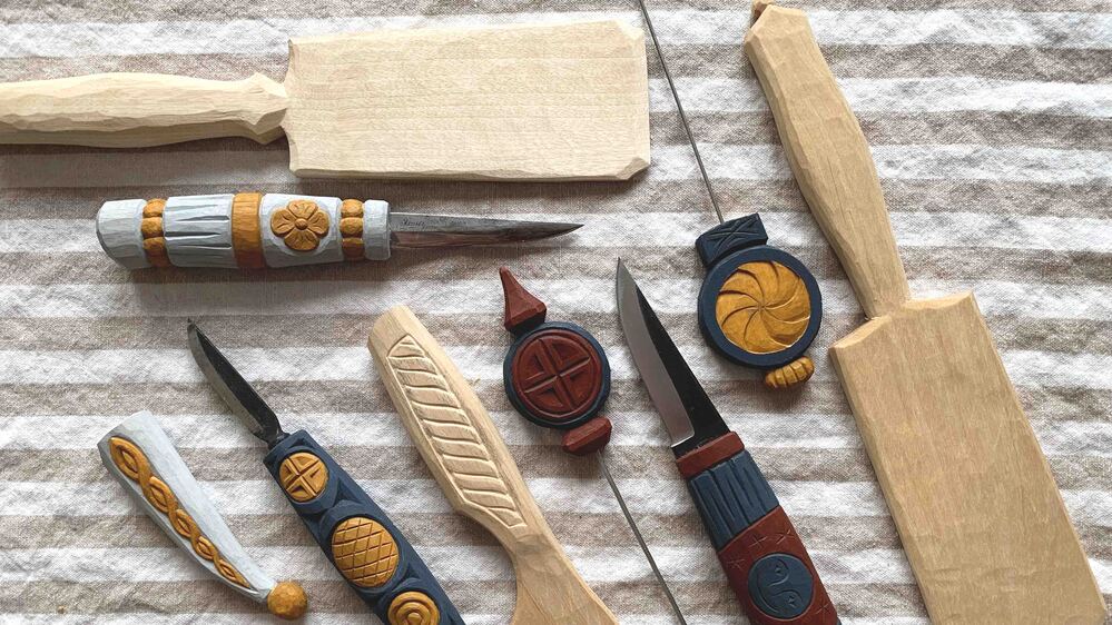 Flera handgjorda träsnideriverktyg och knivar med dekorerade handtag ligger på en randig tygduk.