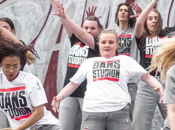 En grupp unga kvinnor dansar energiskt utomhus framför en graffitivägg, alla bär t-shirts med texten 'DANS STUDIO'.