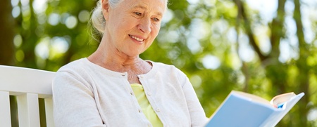 Äldre kvinna med vitt hår sitter utomhus på en bänk och läser en bok, omgiven av grönska.