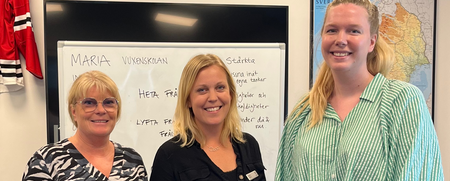 Tre kvinnor står framför en whiteboard i ett klassrum eller mötesrum och ler mot kameran.