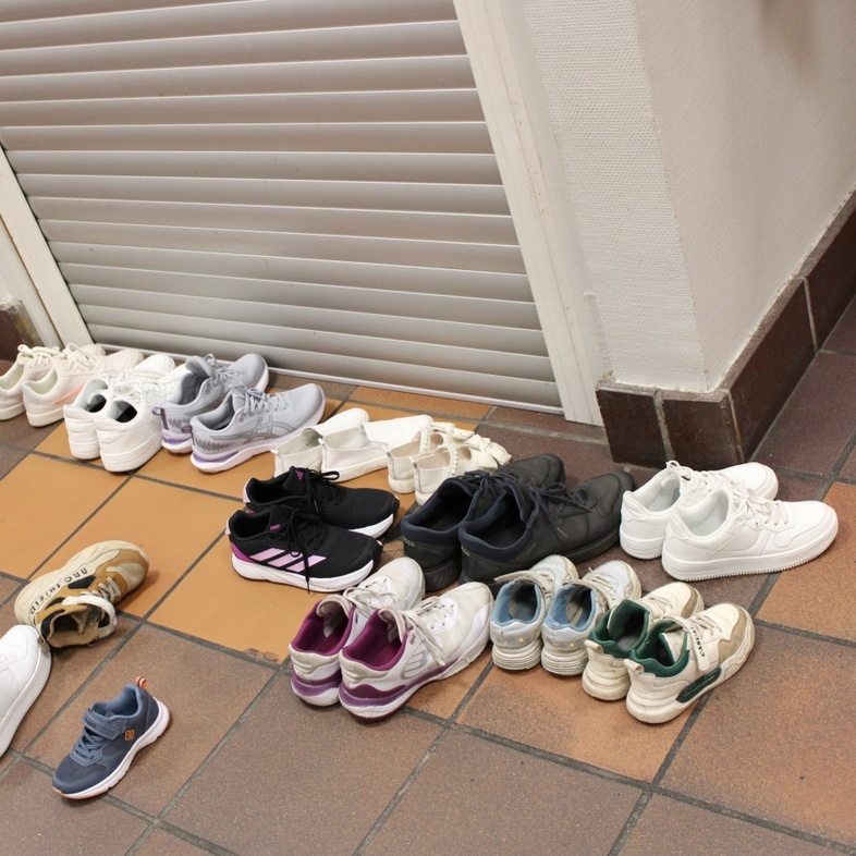 Flera par skor och sandaler står uppradade på ett klinkergolv i en korridor vid en stängd jalusidörr.