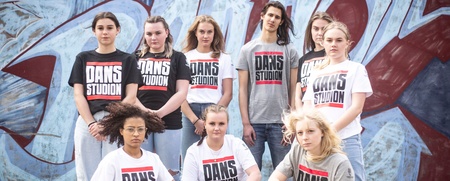En grupp unga personer poserar framför en graffitivägg, alla bär t-shirts med texten 'Dansstudion'.