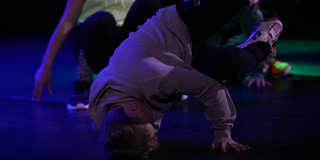 En person utför ett breakdance-move på golvet med stöd av ena handen, andra personer i bakgrunden.