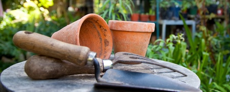 Trädgårdsredskap och lerkrukor på ett bord i en grönskande trädgård med växthus i bakgrunden.