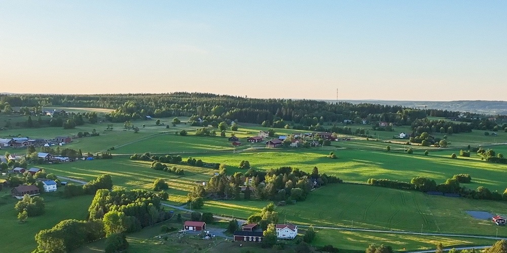 Landskapsvy över gröna fält, skogsdungar och några gårdar under en klar blå himmel.