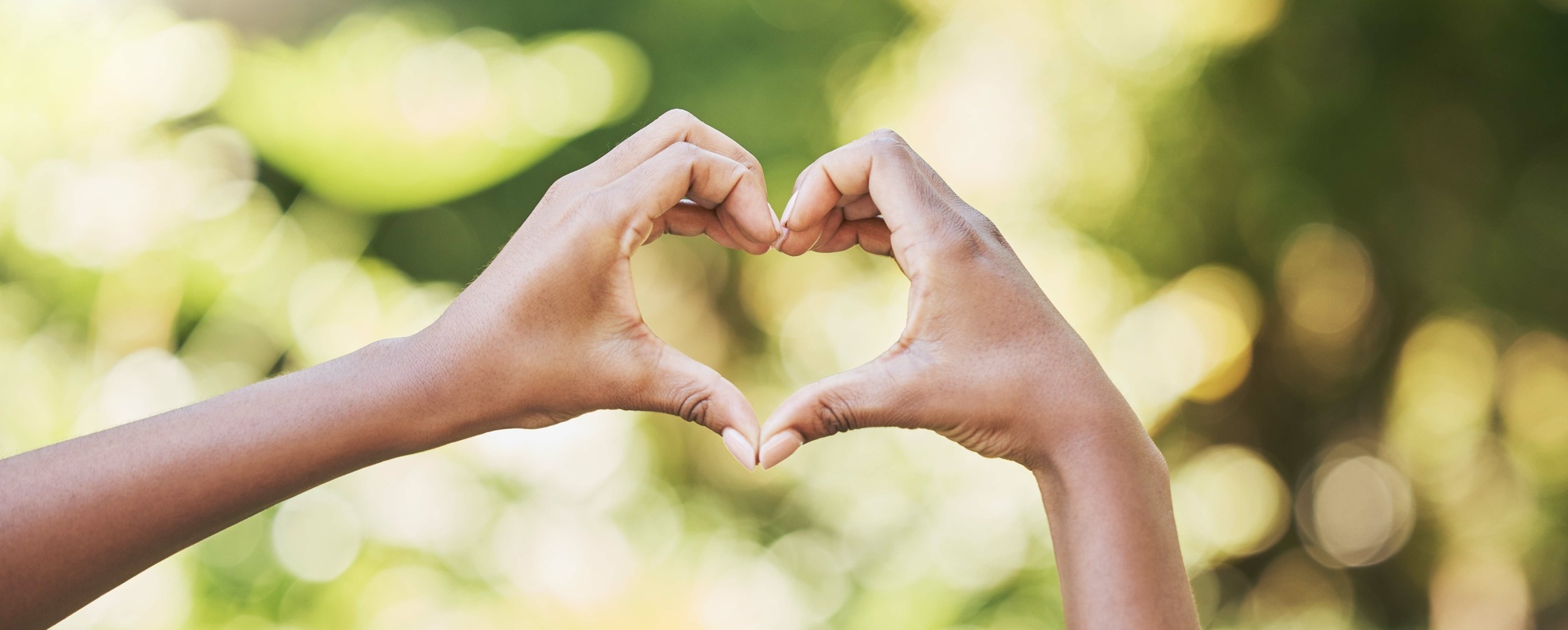 Två händer formar ett hjärta mot en suddig, grön bakgrund utomhus.