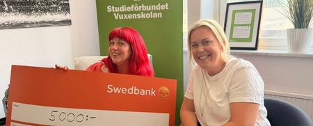 Två personer sitter med en stor orange check från Swedbank på 5000 kronor framför en grön skylt från Vuxenskolan.