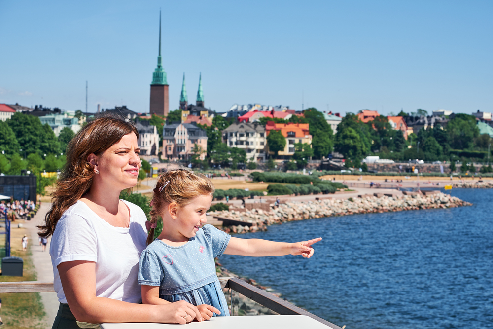 En kvinna och ett barn står vid vattnet och tittar ut över en stad i solsken.