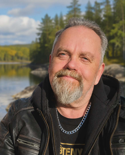 Man med grått skägg och läderjacka poserar utomhus vid en sjö med skog i bakgrunden.