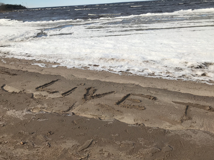 Texten 'LIVET' är skriven i sanden på en strand med vågor och skum i bakgrunden.