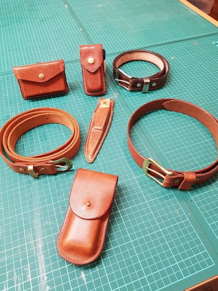 Flera handgjorda läderprodukter, inklusive bälten, fodral och armband, ligger på ett grönt skärmatta.