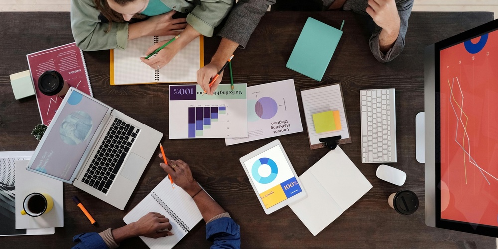 Tre personer arbetar tillsammans vid ett skrivbord med datorer, dokument och diagram.