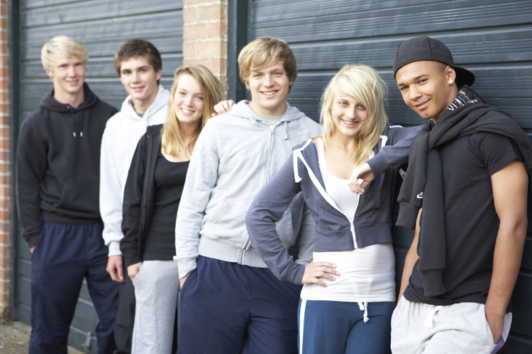 Sex unga personer står avslappnat i rad utomhus framför en blå garageport och ler mot kameran.