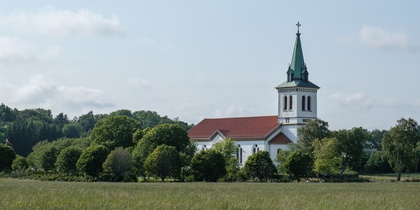En vit kyrka med rött tak och grön spira står omgiven av träd på en äng under blå himmel.