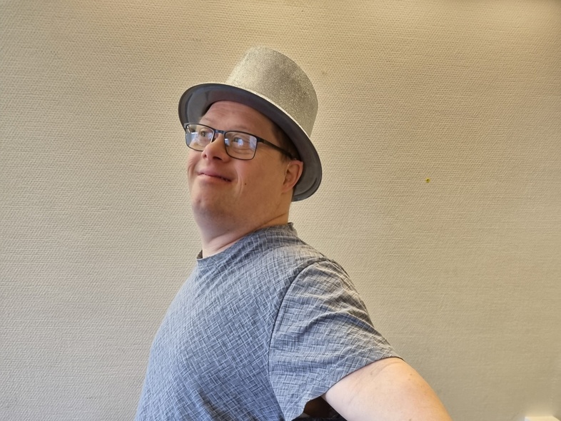 Person med grå hatt och grå t-shirt står med armarna utsträckta mot en ljus vägg.