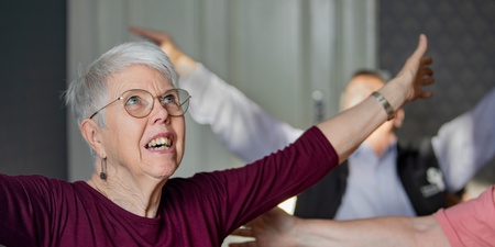 Äldre kvinna med glasögon sträcker ut armarna i ett rum, deltar i gruppaktivitet.