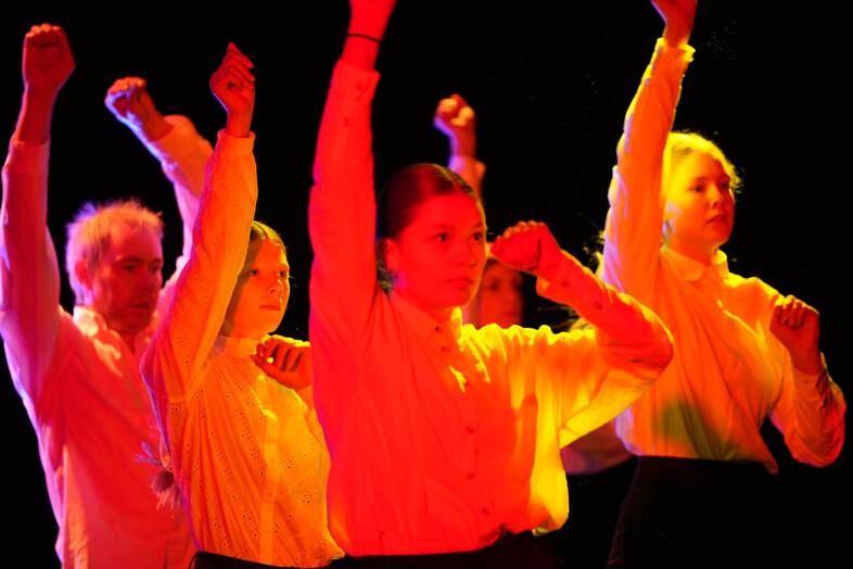 Flera personer i vita skjortor står i rad med armarna upplyfta, belysta av starkt rött och gult scenljus.