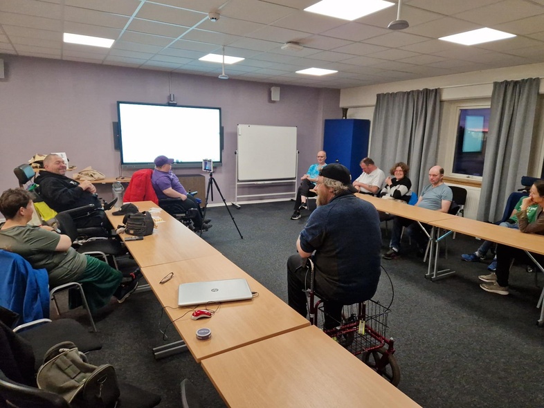 En grupp människor sitter i en U-form runt bord i ett mötesrum med en stor skärm och whiteboard.