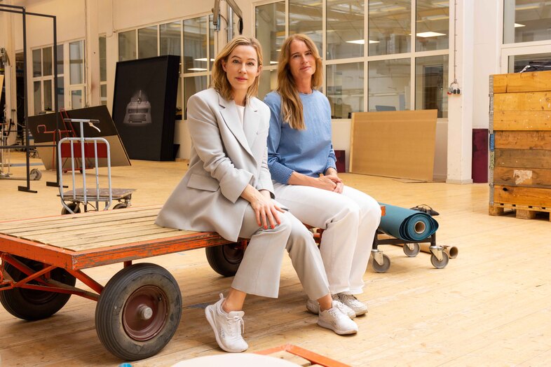 Två kvinnor sitter på en vagn i en ljus industrilokal med trä- och metallföremål runt sig.