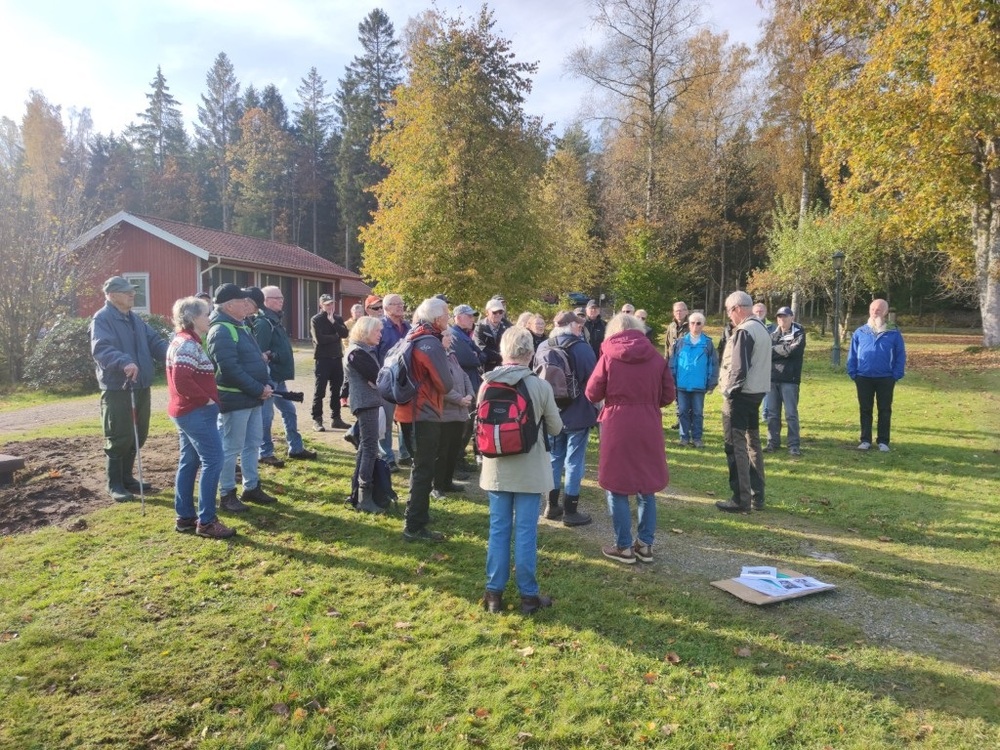 En grupp människor står utomhus på gräs, lyssnar på en guide vid en röd stuga i höstlandskap.