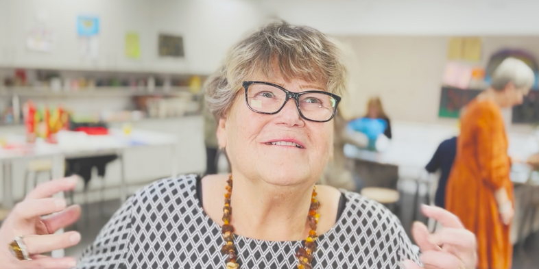 Äldre kvinna med glasögon och halsband gestikulerar i ett ljust rum med andra personer i bakgrunden.