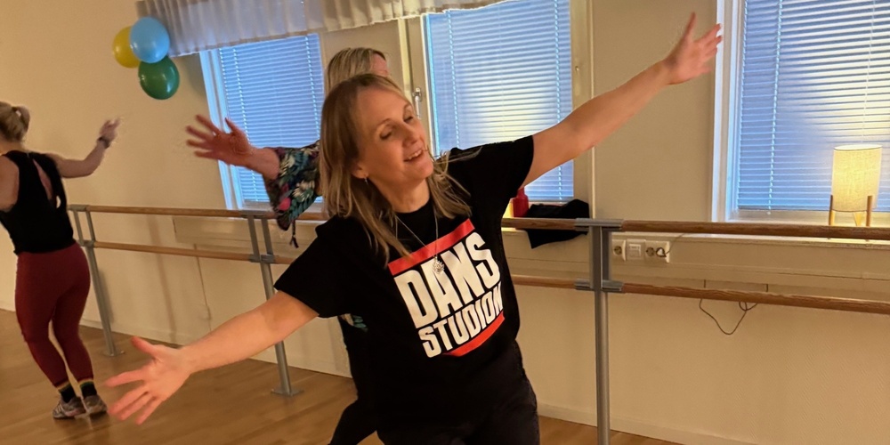 Kvinna dansar glatt i en dansstudio med trägolv och speglar, iförd svart t-shirt med texten 'DANS STUDIO'.