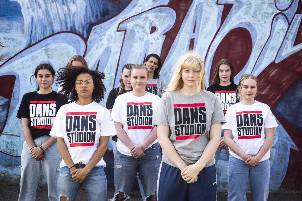 En grupp unga personer står framför en graffitivägg, alla bär t-shirts med texten 'DANS STUDION'.