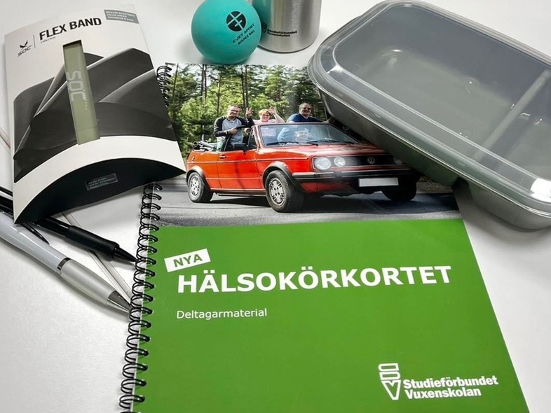 Ett grönt häfte med texten 'Hälsokörkortet', pennor, matlåda, träningsband och broschyr på ett bord.