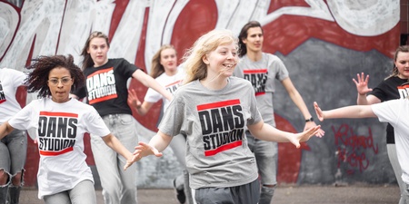 En grupp unga personer dansar energiskt utomhus framför en graffitivägg, alla bär t-shirts med texten 'DANS STUDIO'.