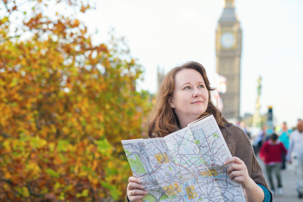 Kvinna håller en karta och står utomhus nära Big Ben i London, med höstlöv i bakgrunden.