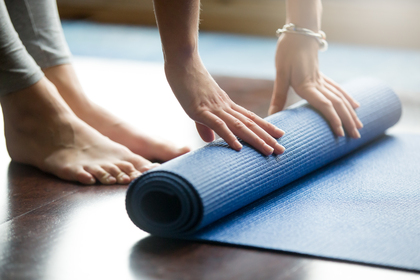 En person rullar ut en blå yogamatta på ett trägolv, bara fötter och händer syns.