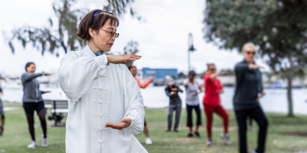 En grupp människor utövar tai chi utomhus i en park, med en kvinna i vit dräkt i förgrunden.