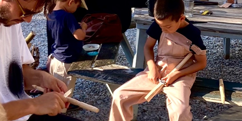 Barn och vuxna sitter utomhus och täljer träpinnar tillsammans vid ett bord.