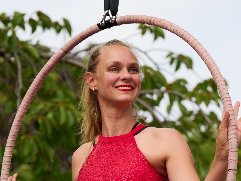 Kvinna i röd topp poserar leende i en cirkusring utomhus med gröna träd i bakgrunden.