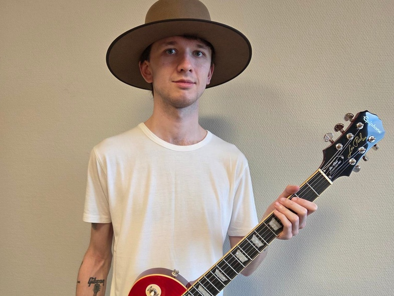 Person med bredbrättad hatt och vit t-shirt håller en röd och gul elgitarr mot en ljus vägg.